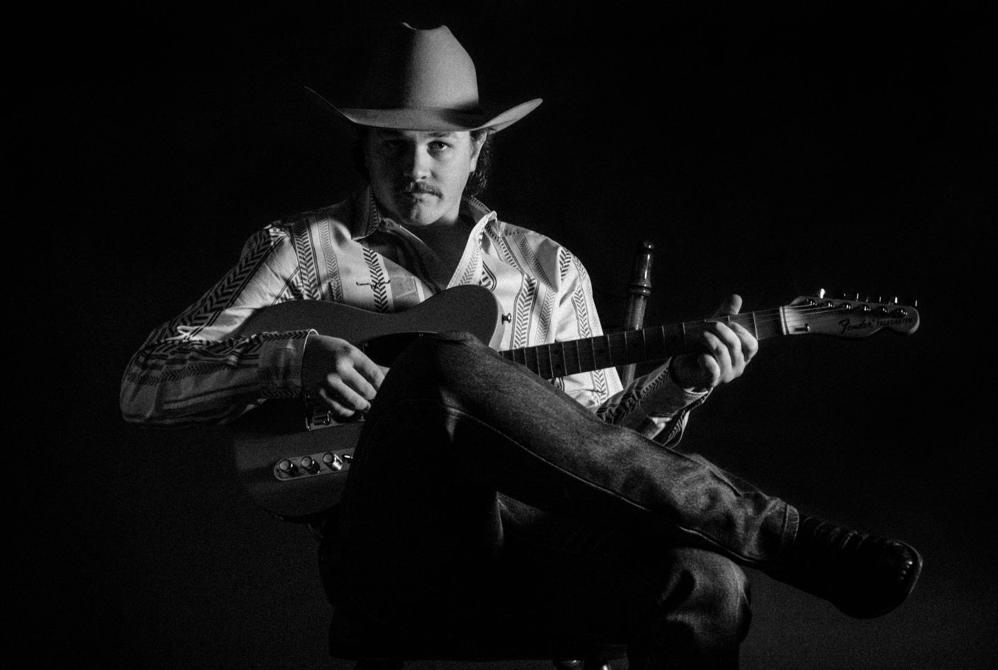Black & white image of a man sat holding a guitar wearing a cowboy hat, shirt, jeans and boots